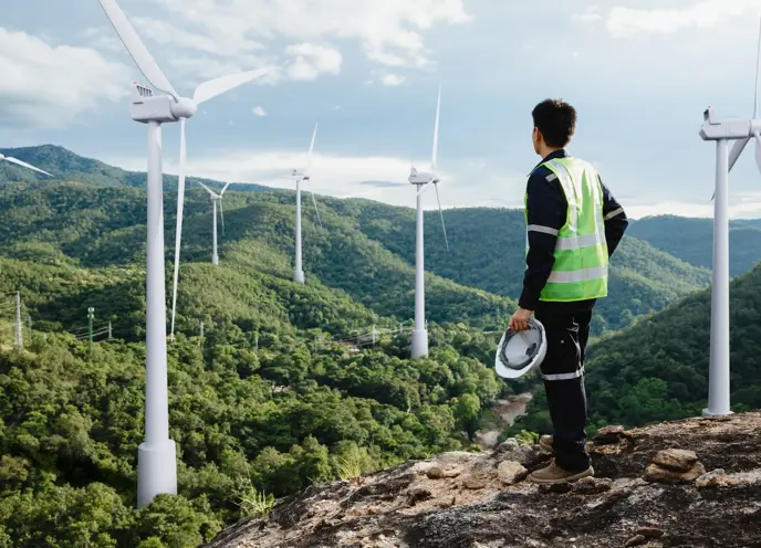 A man stood on a hill top in high-vis holding a hard hat looking out over a forest with wind turbines.